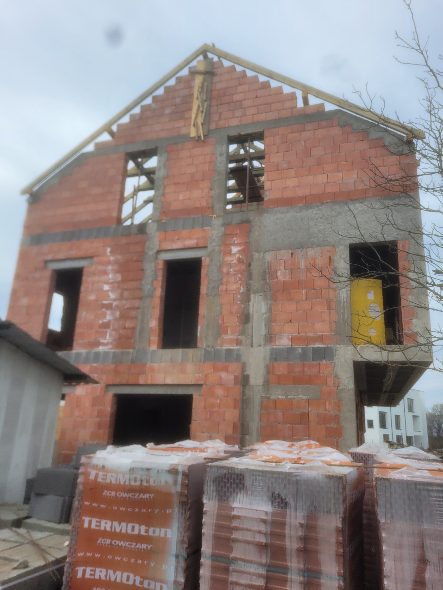 Surowy stan dwupiętrowego budynku z czerwonej cegły w trakcie budowy, z widocznymi otworami okiennymi i niezadaszonym dachem, palety cegieł TERMOTON na pierwszym planie.