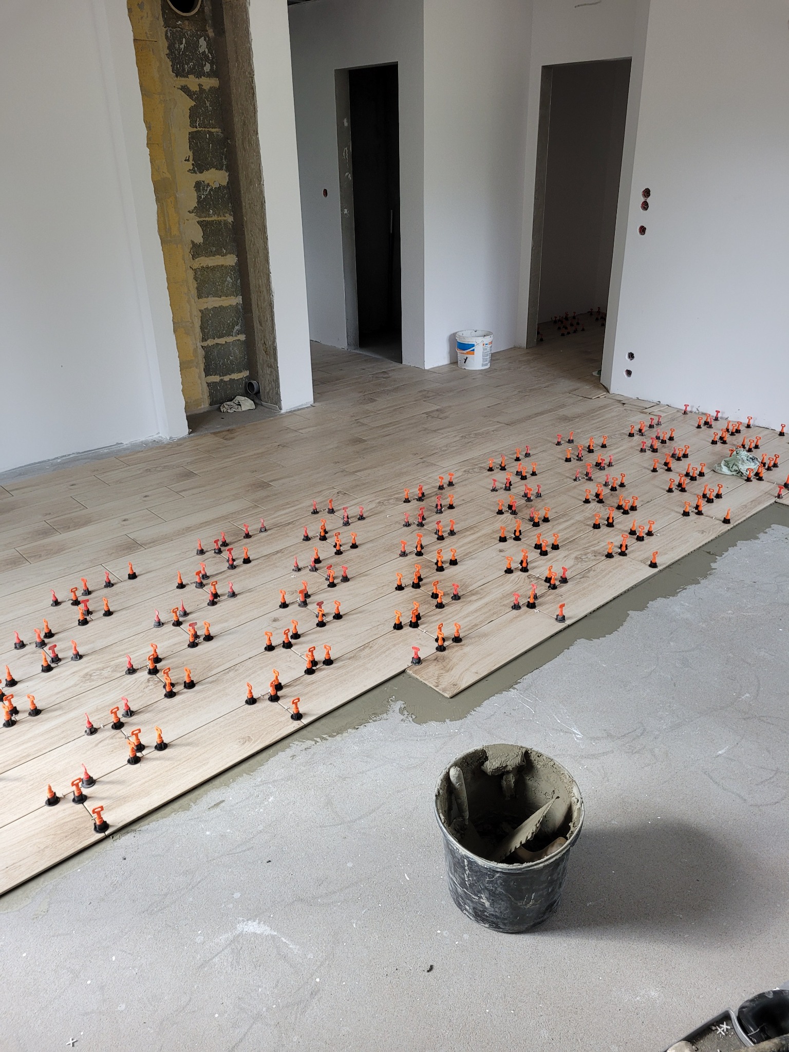 Układanie podłogi z płytek imitujących drewno z użyciem systemu poziomowania, widoczny kubeł z zaprawą i szpachelką, surowy stan ścian w tle.