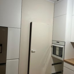 Colorview - Wnętrze nowocześnie urządzonej kuchni w trakcie wykańczania, widoczne białe szafki kuchenne, lodówka, piekarnik oraz ściana w odcieniach beżu z ukrytymi drzwiami, na podłodze resztki materiałów...