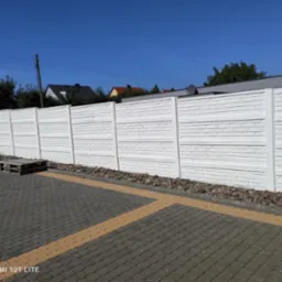 Gotowy, pomalowany na biało prefabrykowany betonowy płot o strukturze cegły, oddzielający posesję od chodnika wyłożonego kostką brukową w odcieniach szarości i beżu.