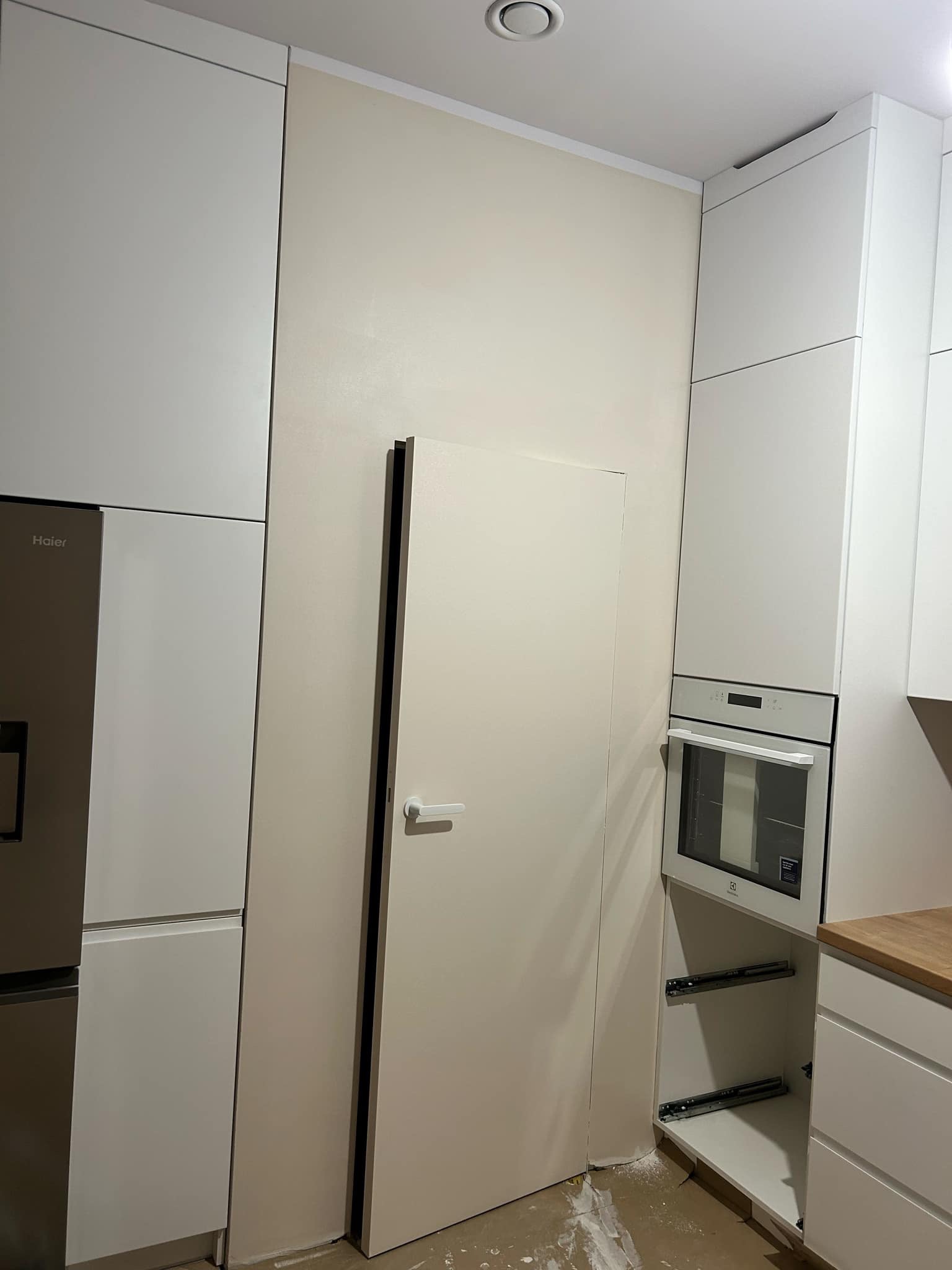 Wnętrze nowocześnie urządzonej kuchni w trakcie wykańczania, widoczne białe szafki kuchenne, lodówka, piekarnik oraz ściana w odcieniach beżu z ukrytymi drzwiami, na podłodze resztki materiałów...