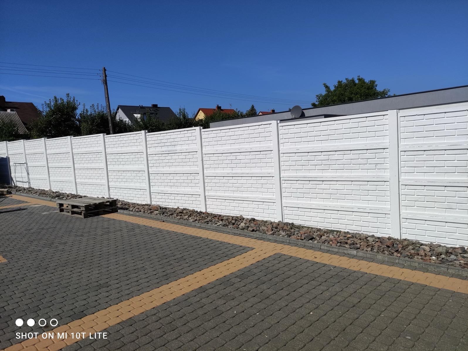 Gotowy, pomalowany na biało prefabrykowany betonowy płot o strukturze cegły, oddzielający posesję od chodnika wyłożonego kostką brukową w odcieniach szarości i beżu.