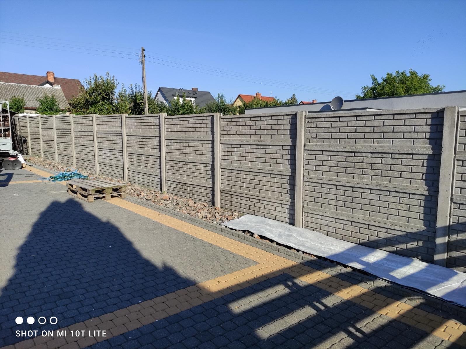 Długi, prefabrykowany betonowy płot z elementami imitującymi cegłę, oddzielający posesję, z drewnianą paletą i niebieskim przewodem na brukowanym podjeździe w słoneczny dzień.