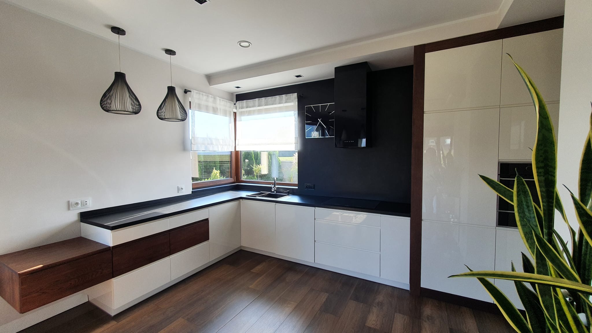Nowoczesna kuchnia z białymi szafkami, ciemnym blatem i drewnianymi akcentami, oświetlona wiszącymi lampami i naturalnym światłem z okna.