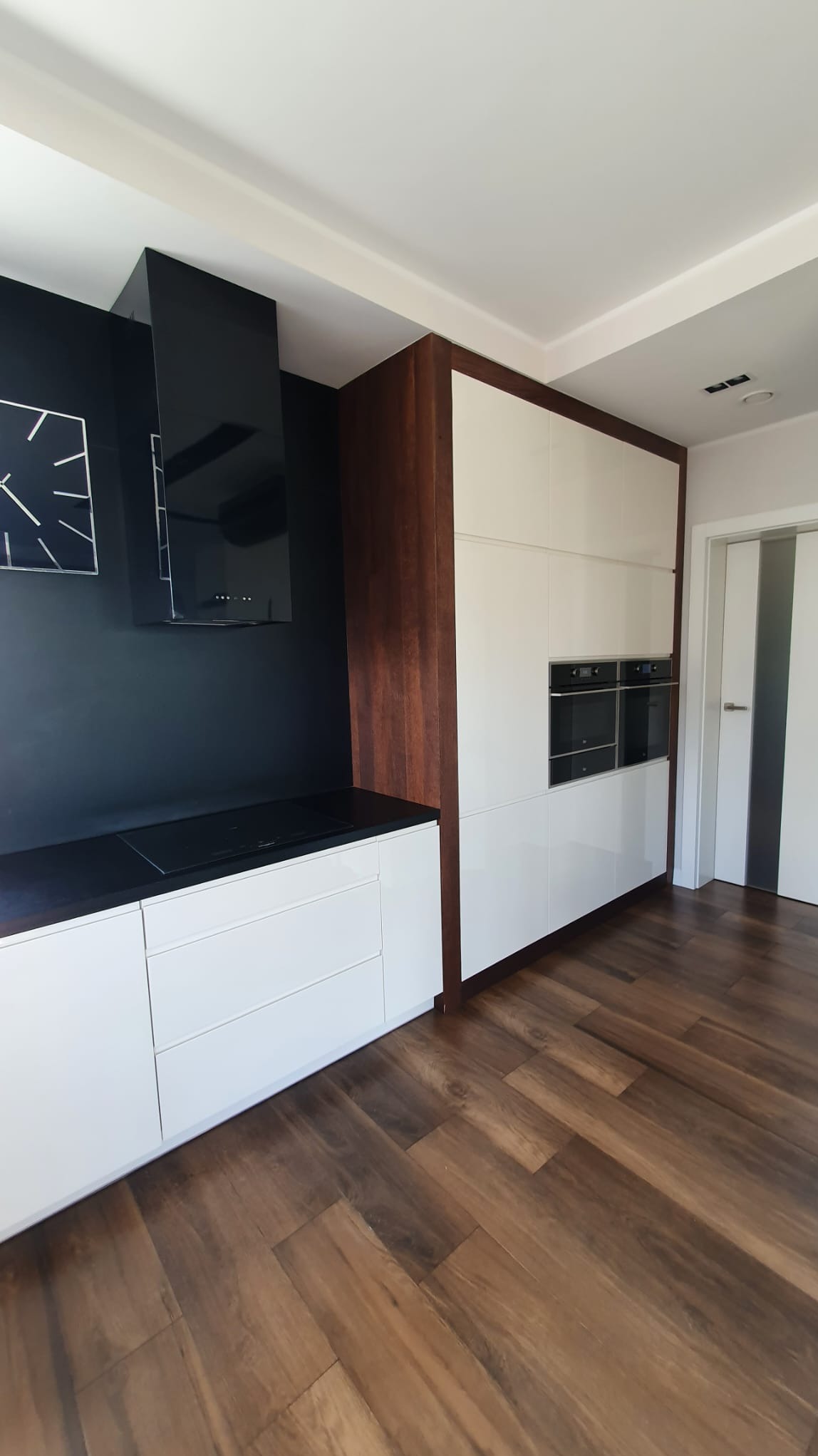 Nowoczesna kuchnia w Stargardzie z białymi szafkami, czarnym blatem i okapem, brązowe wykończenie szafek i drewnopodobna podłoga ułożona w jodełkę.