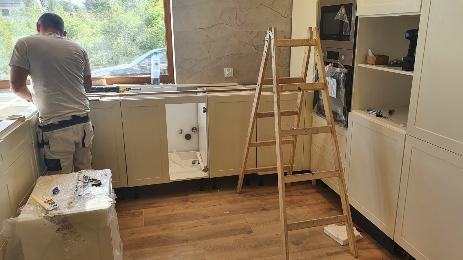 Człowiek w roboczych spodniach montuje jasne szafki kuchenne, w tle widoczne okno i drabina.
