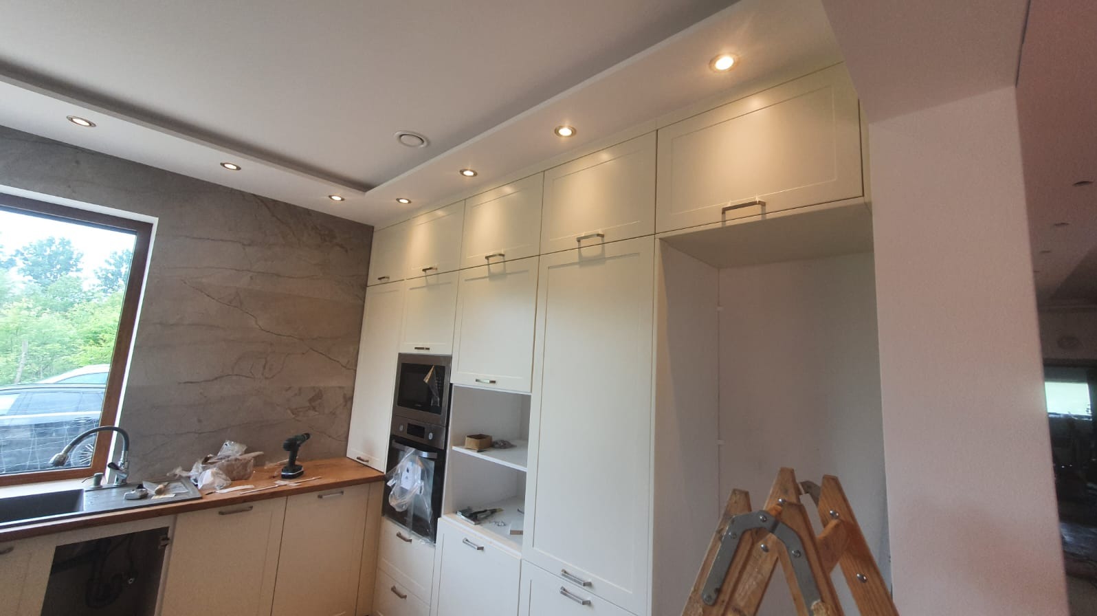 Wnętrze kuchni w trakcie montażu zabudowy meblowej z drewnianą drabiną i narzędziami widocznymi na blacie.