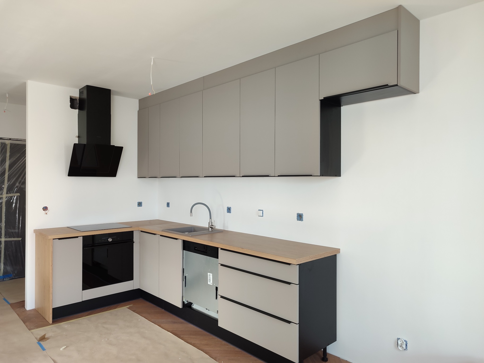 Nowoczesna kuchnia w odcieniach szarości z drewnianym blatem, czarnym okapem i niedokończonymi detalami instalacji elektrycznej na białej ścianie.