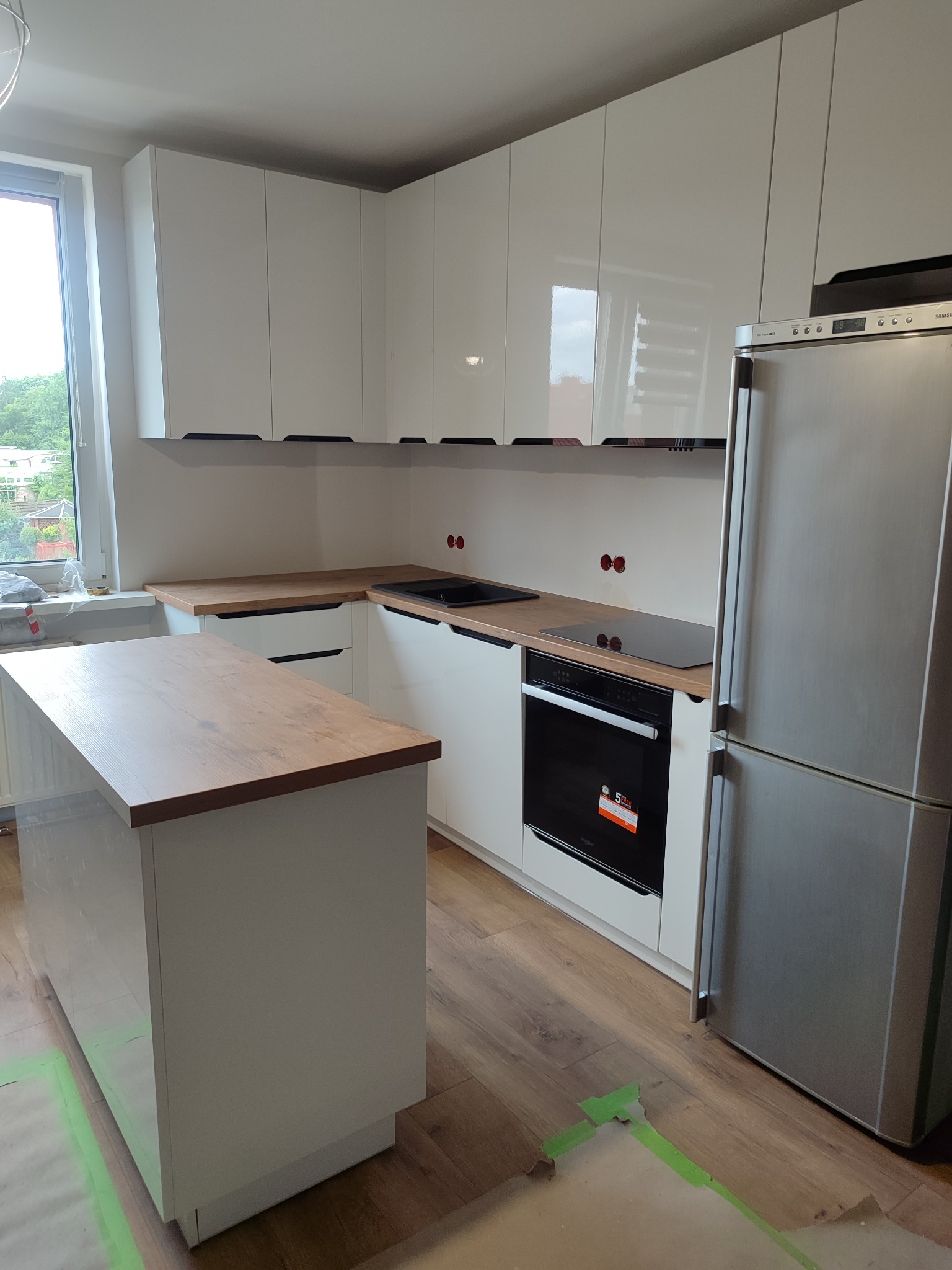 Nowoczesna, jasna kuchnia z białymi, błyszczącymi szafkami i drewnianym blatem. Widoczna wyspa kuchenna i sprzęt AGD. Podłoga zabezpieczona taśmą malarską.