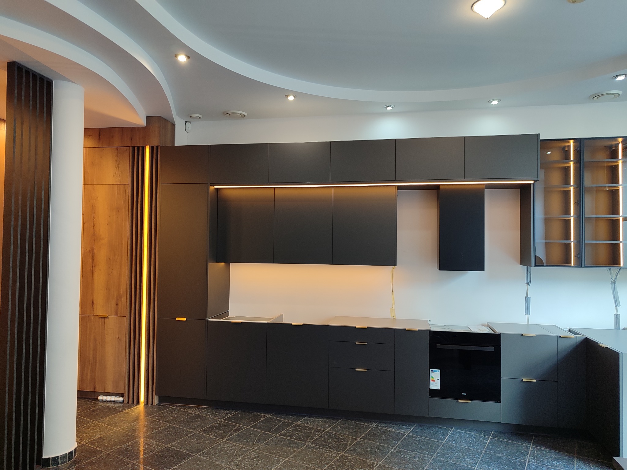 Nowoczesna kuchnia z ciemnymi, matowymi frontami, oświetleniem LED i drewnianymi akcentami. Widok na całą zabudowę, od podłogi po sufit.