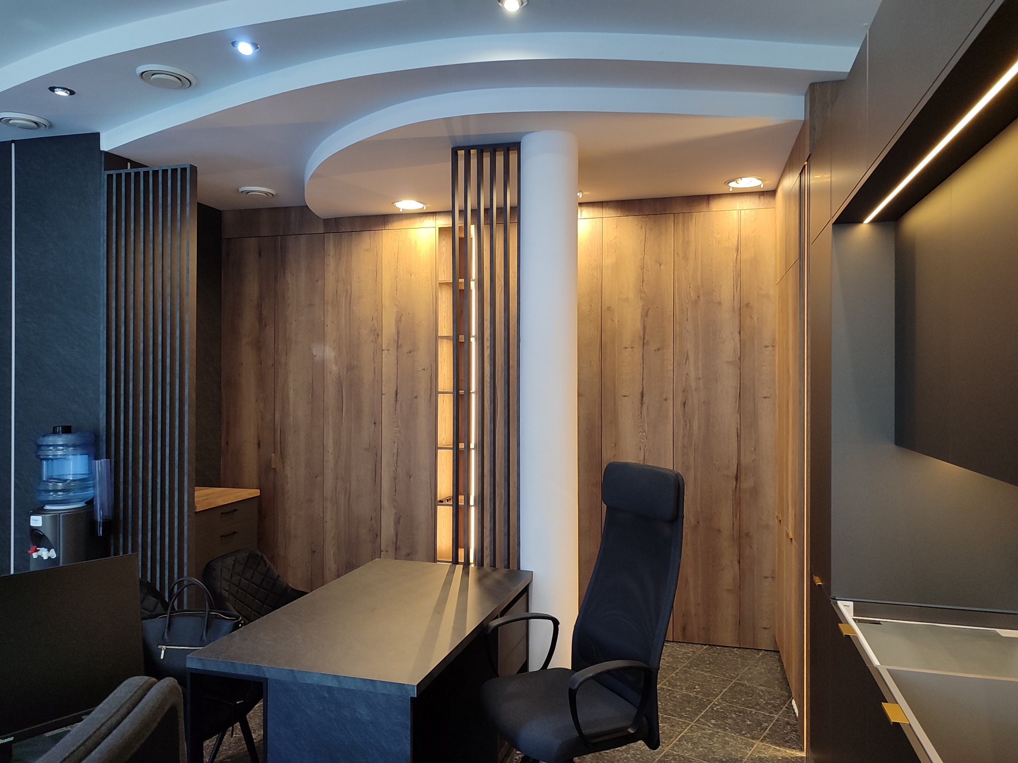 Nowoczesne biuro z drewnianą zabudową, oświetleniem LED i minimalistycznym biurkiem. Widoczny dystrybutor wody i krzesło biurowe.