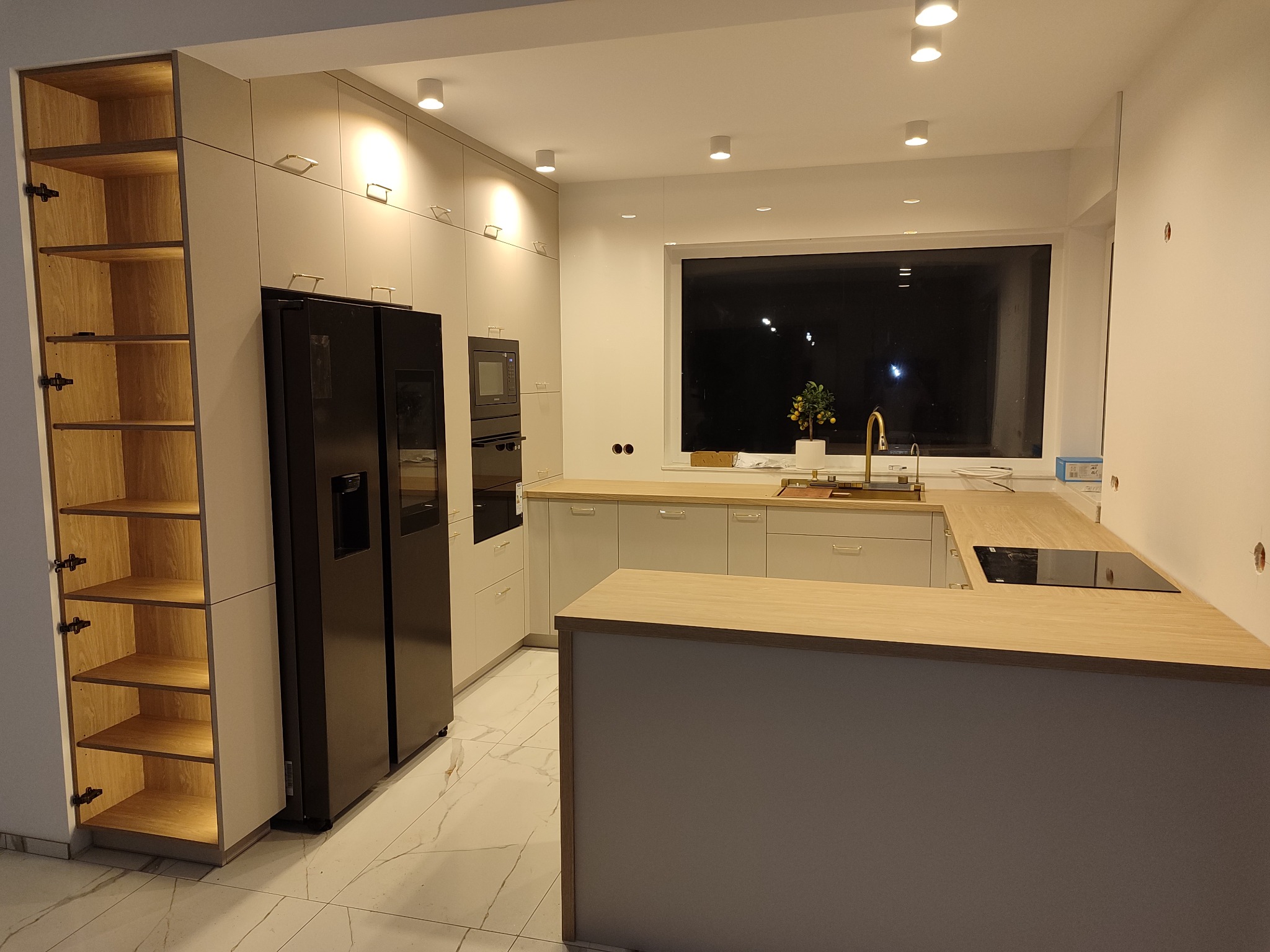 Jasna, nowoczesna kuchnia z wyspą i czarną lodówką side-by-side. Otwarty regał z oświetleniem wewnętrznym po lewej. Okno z widokiem na nocne niebo.