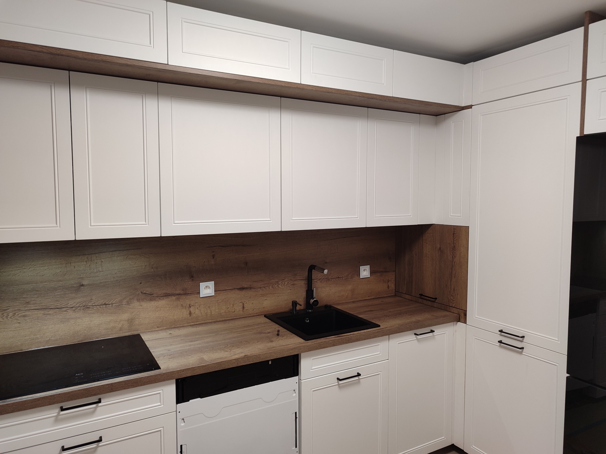 Nowoczesna kuchnia z białymi szafkami i blatem imitującym drewno. Czarny zlew i bateria dodają kontrastu. Widoczna płyta indukcyjna i front zmywarki.