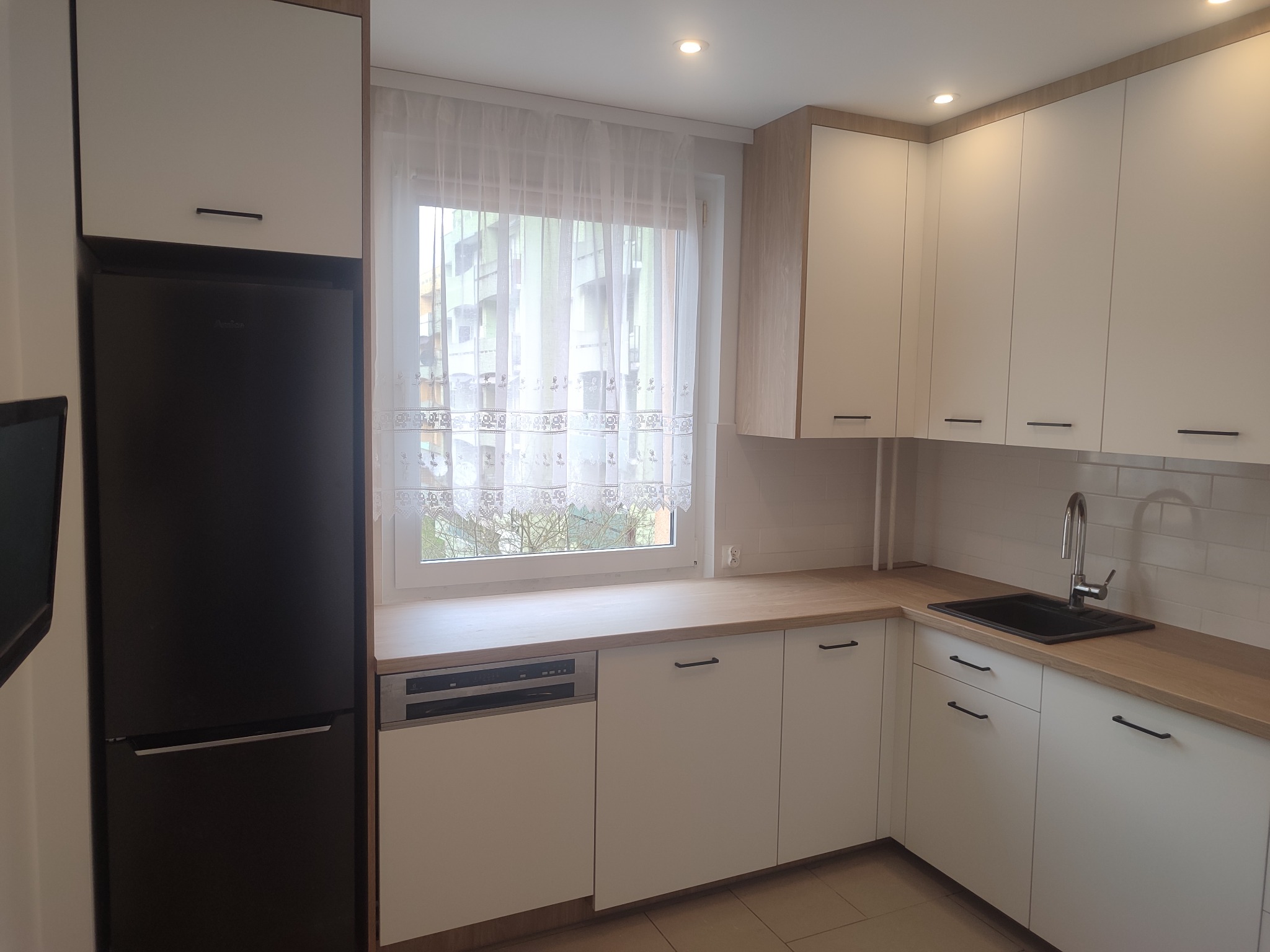 Jasna kuchnia z białymi frontami i drewnianym blatem, czarny zlew i lodówka. Okno z firanką. Minimalistyczny design, nowoczesne wykończenie, oświetlenie sufitowe.