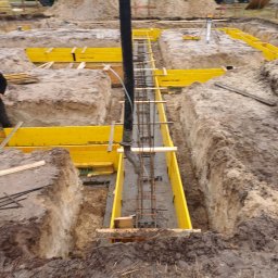 KRAMBUD Dominik Kramek - Wylewanie betonu do wykopu z żółtym szalunkiem i zbrojeniem; w tle betonomieszarki i pracownicy w odblaskowych kamizelkach.