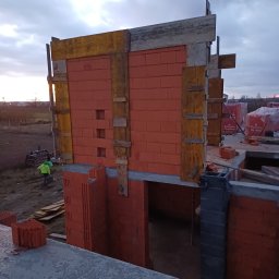 KRAMBUD Dominik Kramek - Wznoszenie ścian domu z czerwonej cegły Porotherm, widoczne drewniane szalunki, na zewnątrz pracownik w odblaskowej kamizelce i palety z materiałem budowlanym, pochmurne niebo w tle.