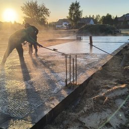 KRAMBUD Dominik Kramek - Wylewanie fundamentów domu jednorodzinnego o wschodzie słońca; robotnicy rozprowadzają mokry beton na przygotowanym zbrojeniu i szalunku.