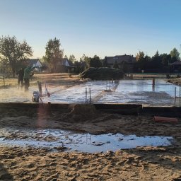 KRAMBUD Dominik Kramek - Wylewanie fundamentów pod budowę domu jednorodzinnego, widoczne zbrojenia i pracownicy w trakcie rozprowadzania betonu, w tle zabudowa jednorodzinna i wschodzące słońce.