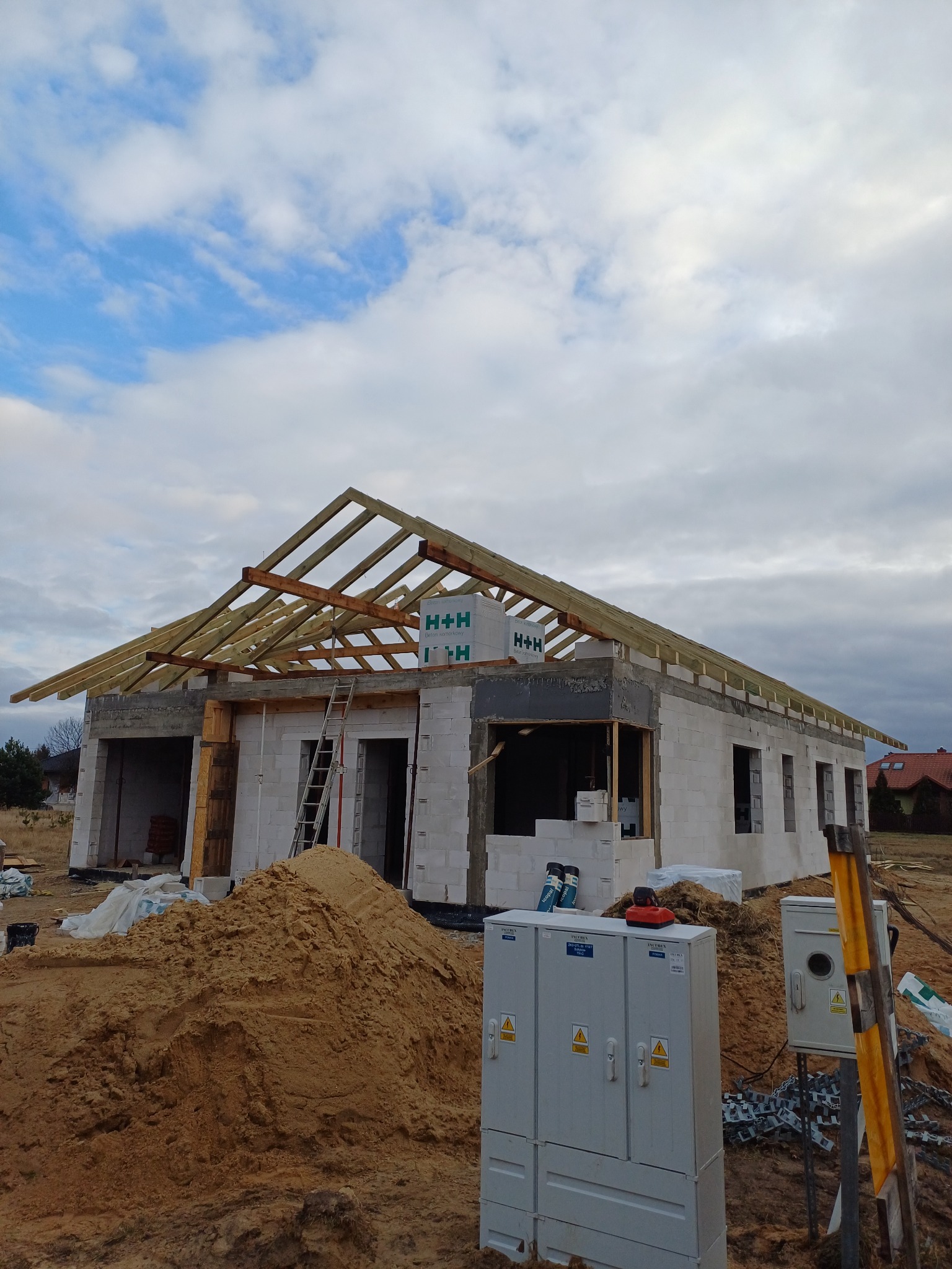 Niedokończony dom z białych bloczków z drewnianą konstrukcją dachu, widoczny stos piasku, skrzynki elektryczne i drabina oparta o ścianę, w tle pochmurne niebo.