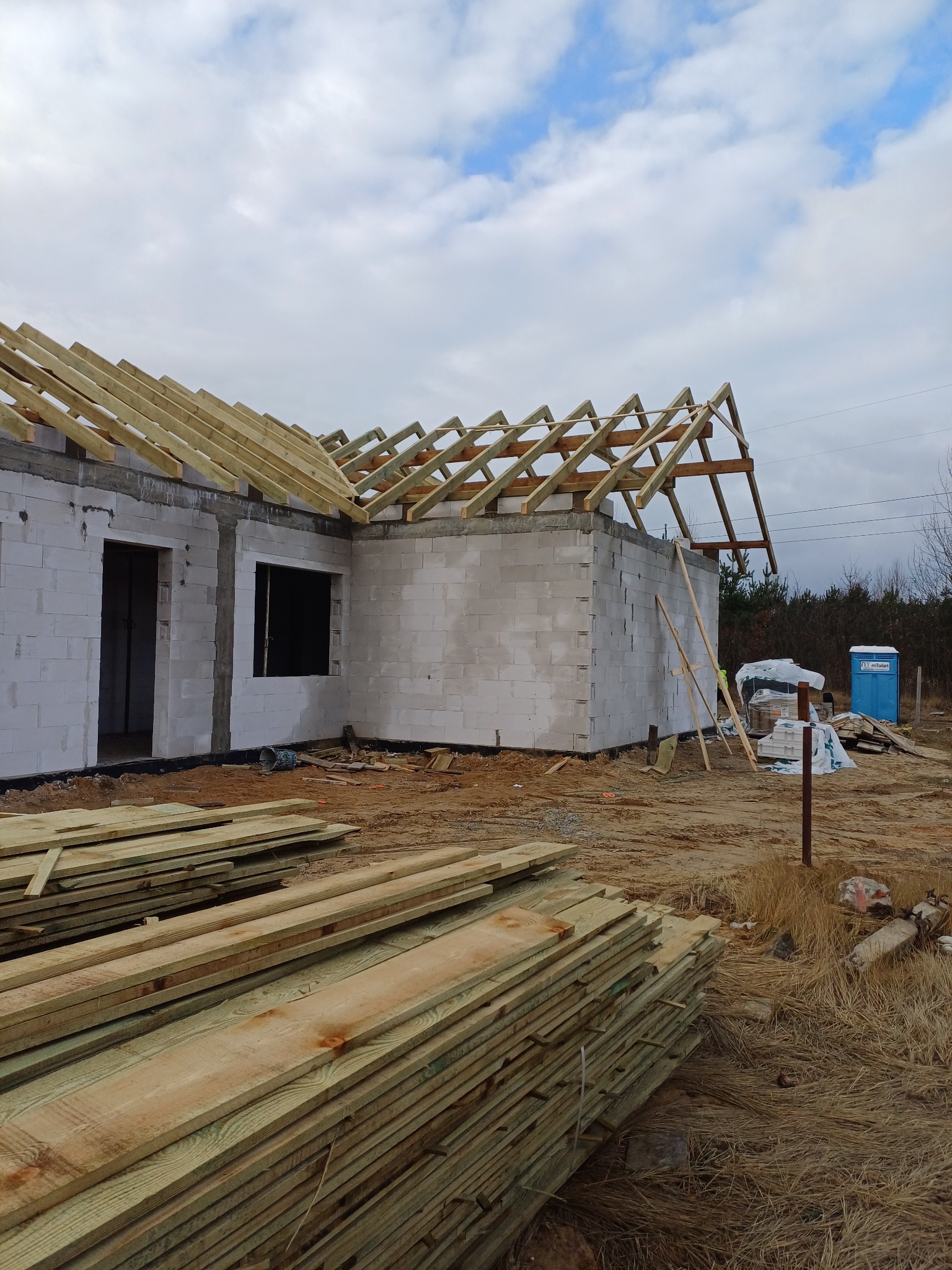 Budowa domu murowanego w trakcie: ściany z bloczków, drewniana konstrukcja dachu, deski na pierwszym planie, przenośna toaleta w tle.