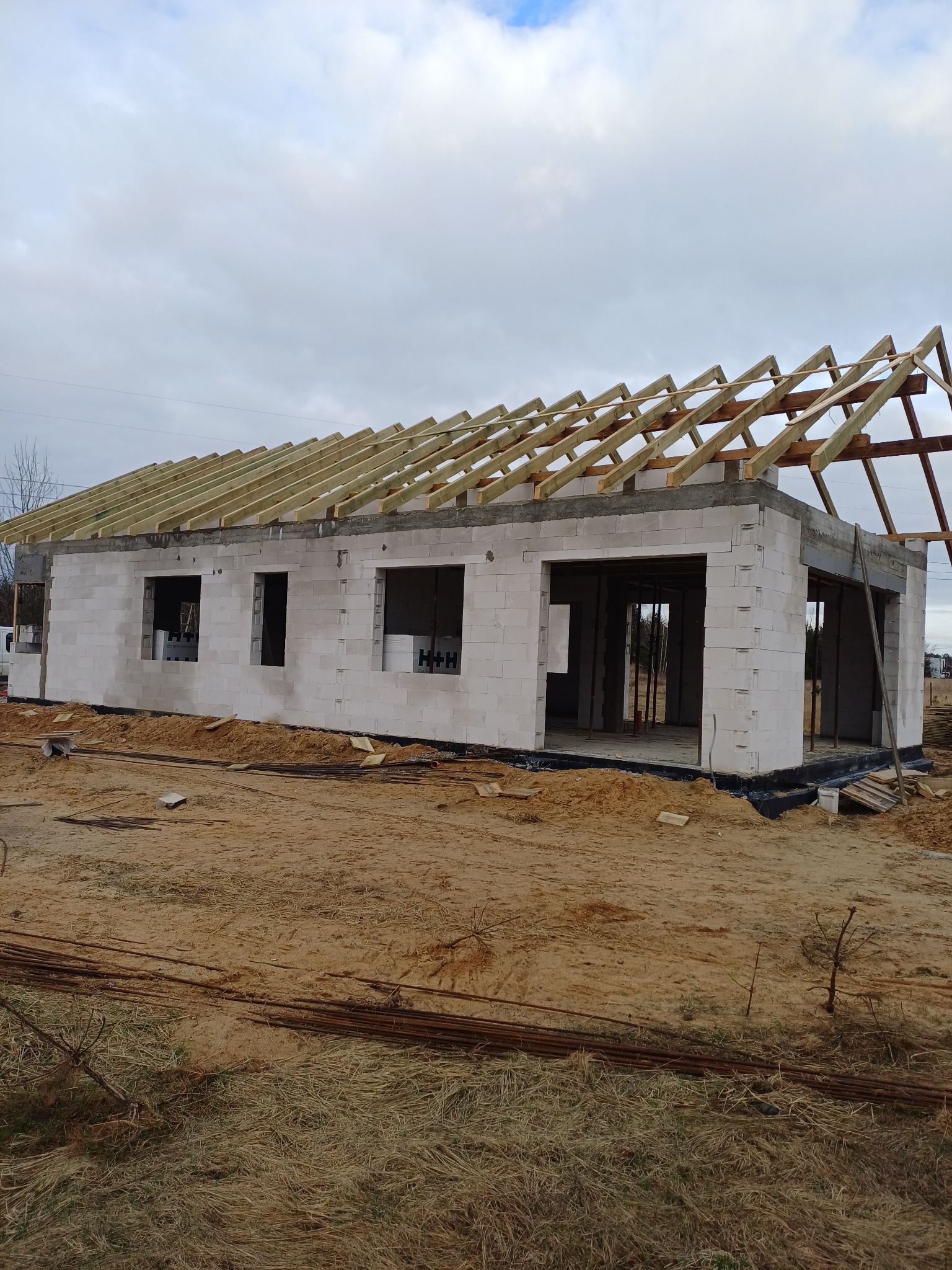 Budowa domu: surowy stan z białych bloczków, widoczne otwory okienne i drzwiowe, konstrukcja drewniana dachu w trakcie montażu, teren wokół w stanie budowy.