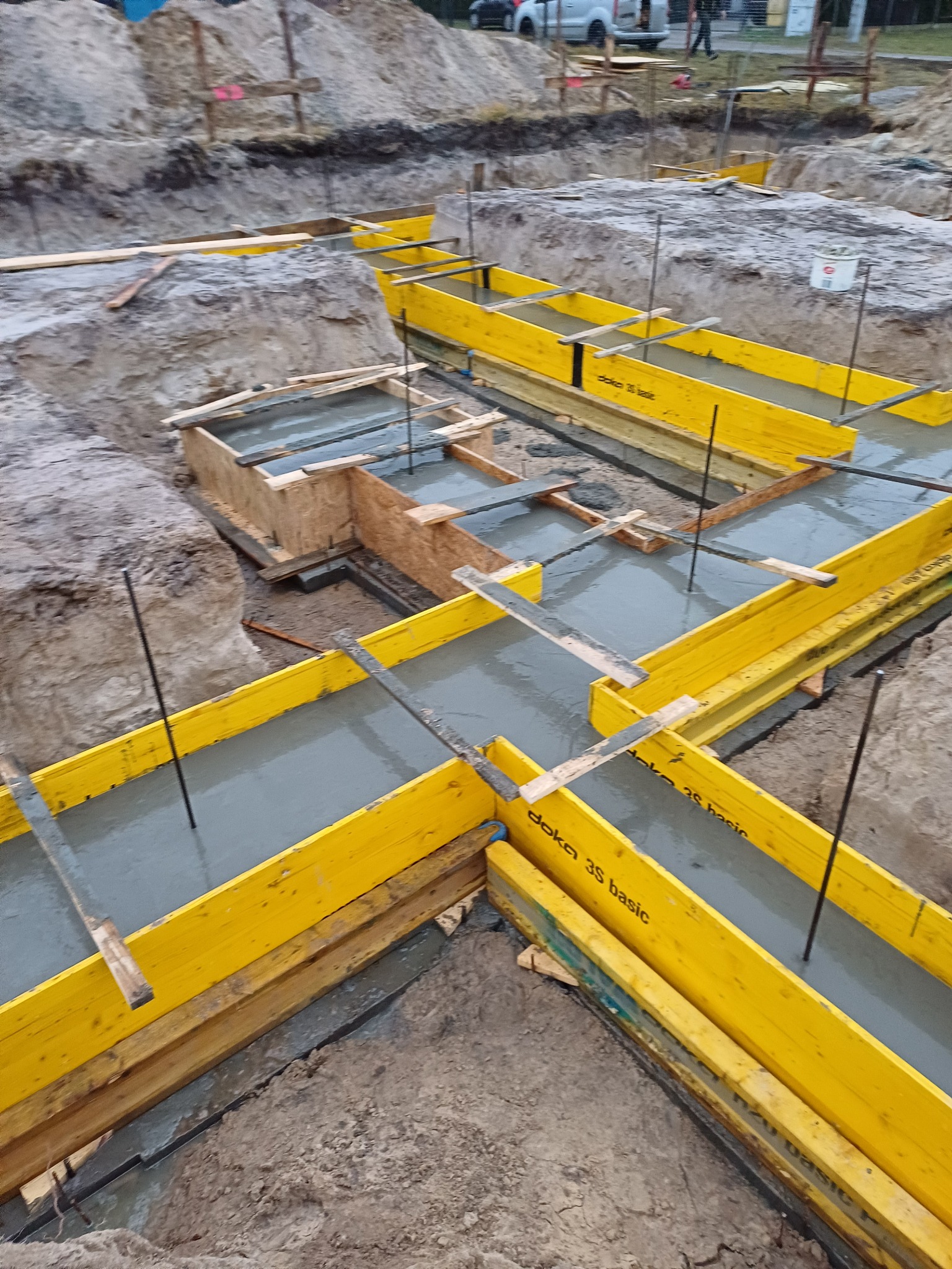 Zalewanie betonem szalunków fundamentowych z żółtymi elementami Doka 3S basic, widoczne wystające pręty zbrojeniowe i wykopany grunt.