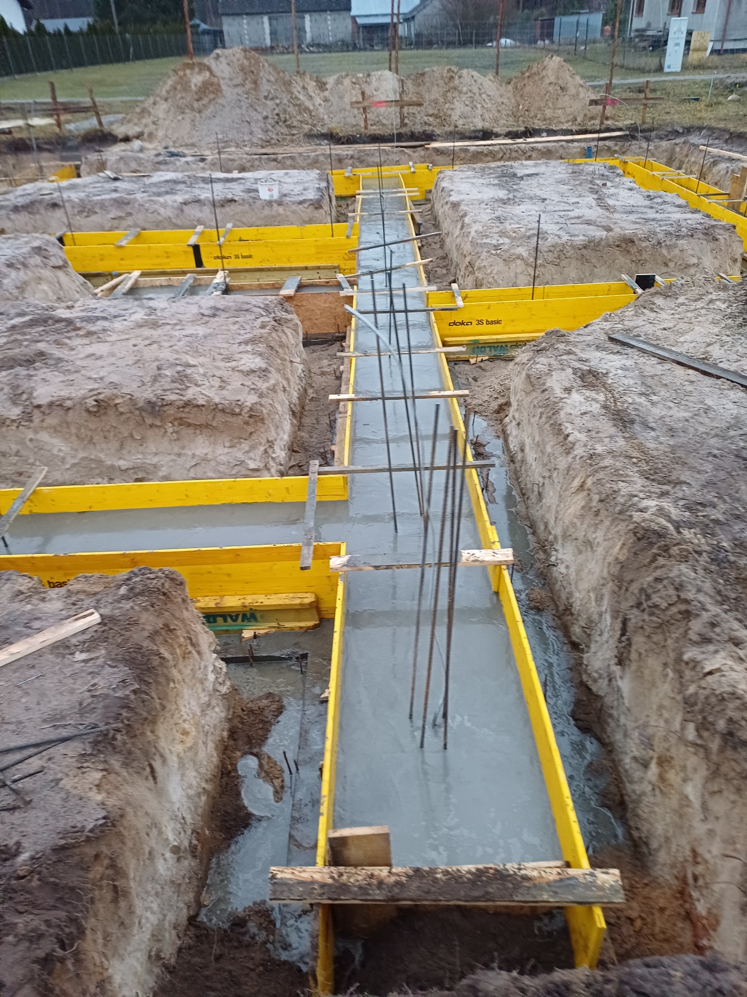 Wykopane fundamenty z żółtym szalunkiem i zbrojeniem zalewane świeżym betonem, widoczne pryzmy ziemi w tle.