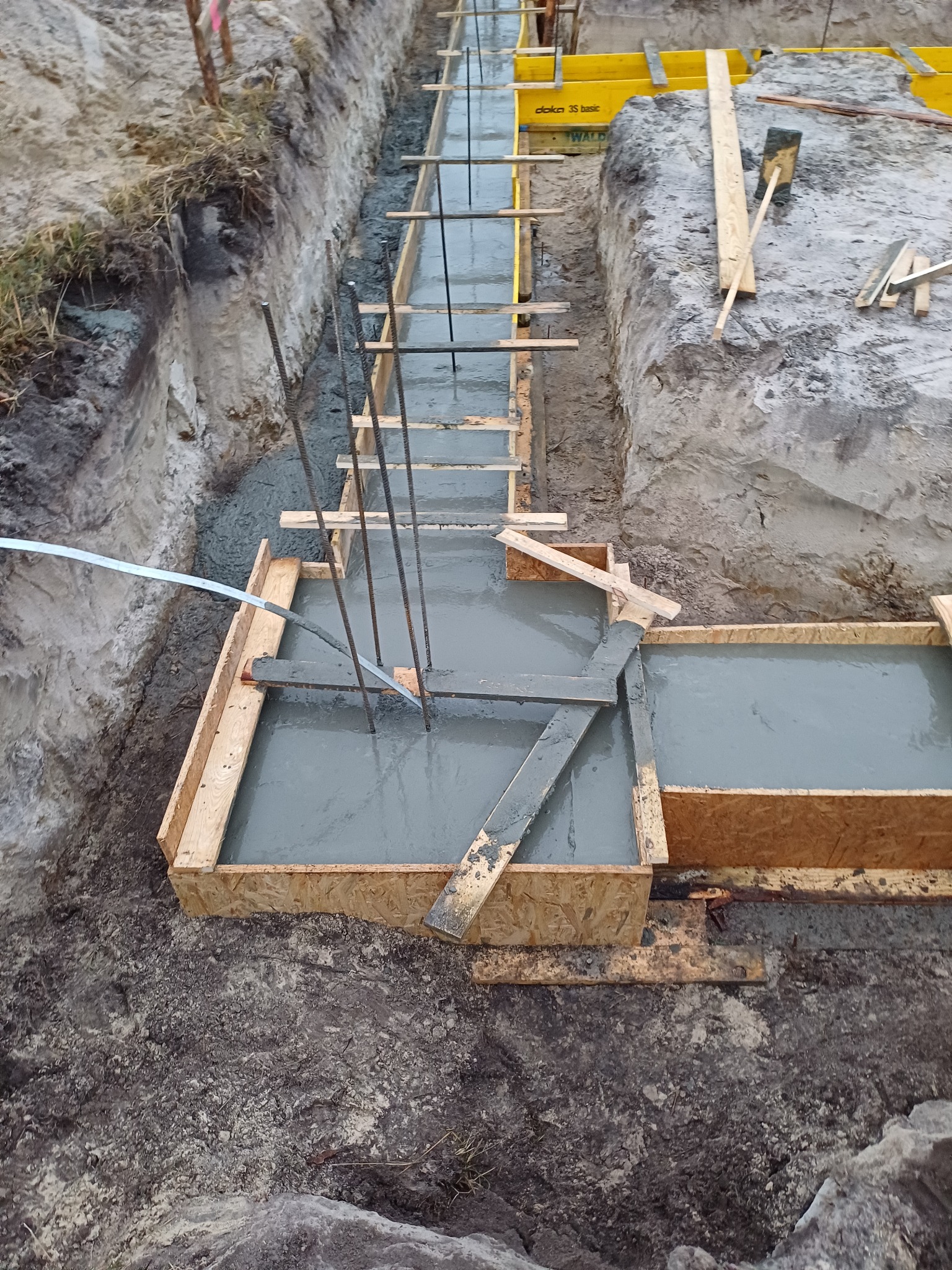 Wykop pod fundamenty zalewany świeżym betonem, z widocznym szalunkiem z desek i wystającymi prętami zbrojeniowymi.