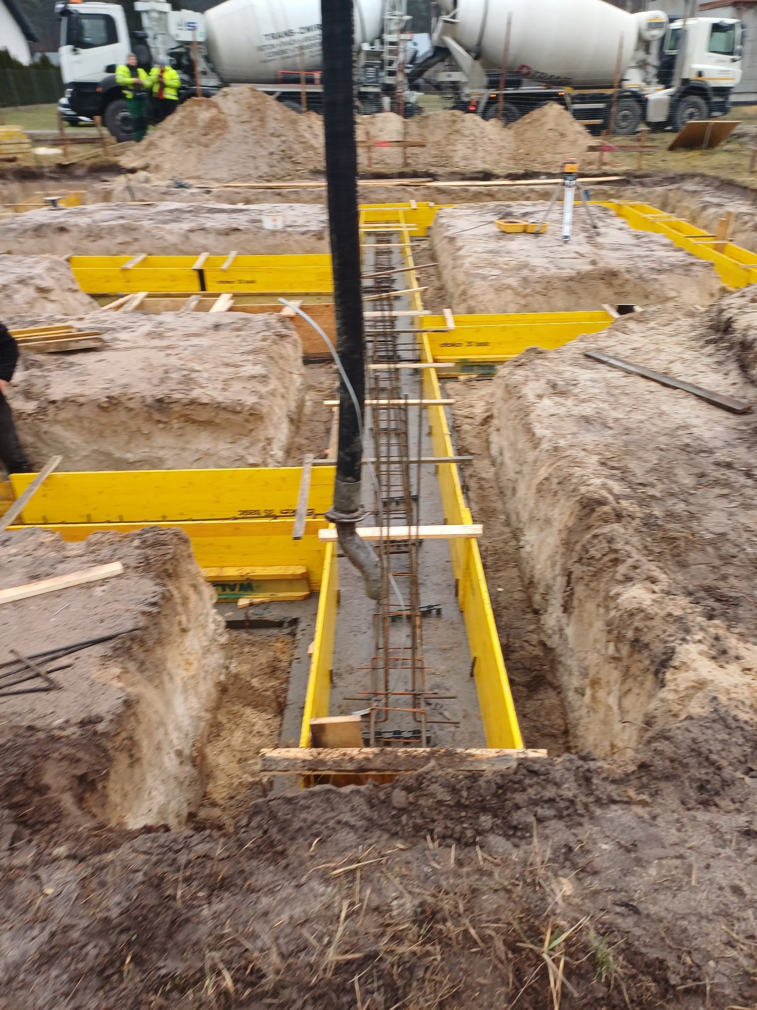 Wylewanie betonu do wykopu z żółtym szalunkiem i zbrojeniem; w tle betonomieszarki i pracownicy w odblaskowych kamizelkach.
