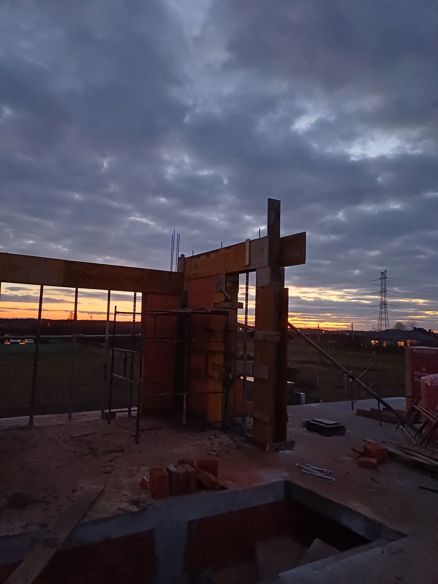 Wczesny etap budowy domu murowanego z widocznym szalunkiem, zbrojeniami i elementami fundamentów na tle zachodzącego słońca i pochmurnego nieba.