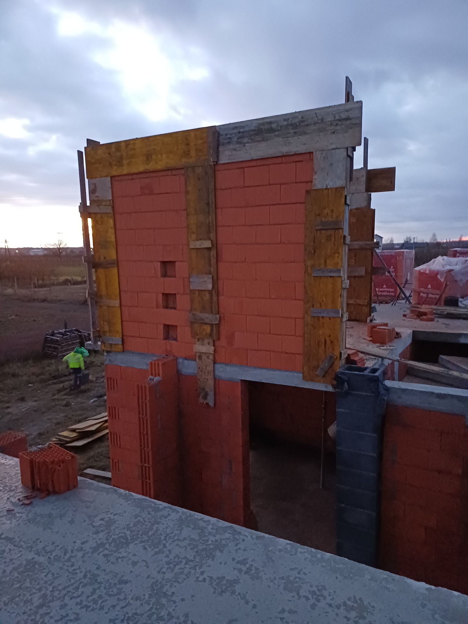 Wznoszenie ścian domu z czerwonej cegły Porotherm, widoczne drewniane szalunki, na zewnątrz pracownik w odblaskowej kamizelce i palety z materiałem budowlanym, pochmurne niebo w tle.