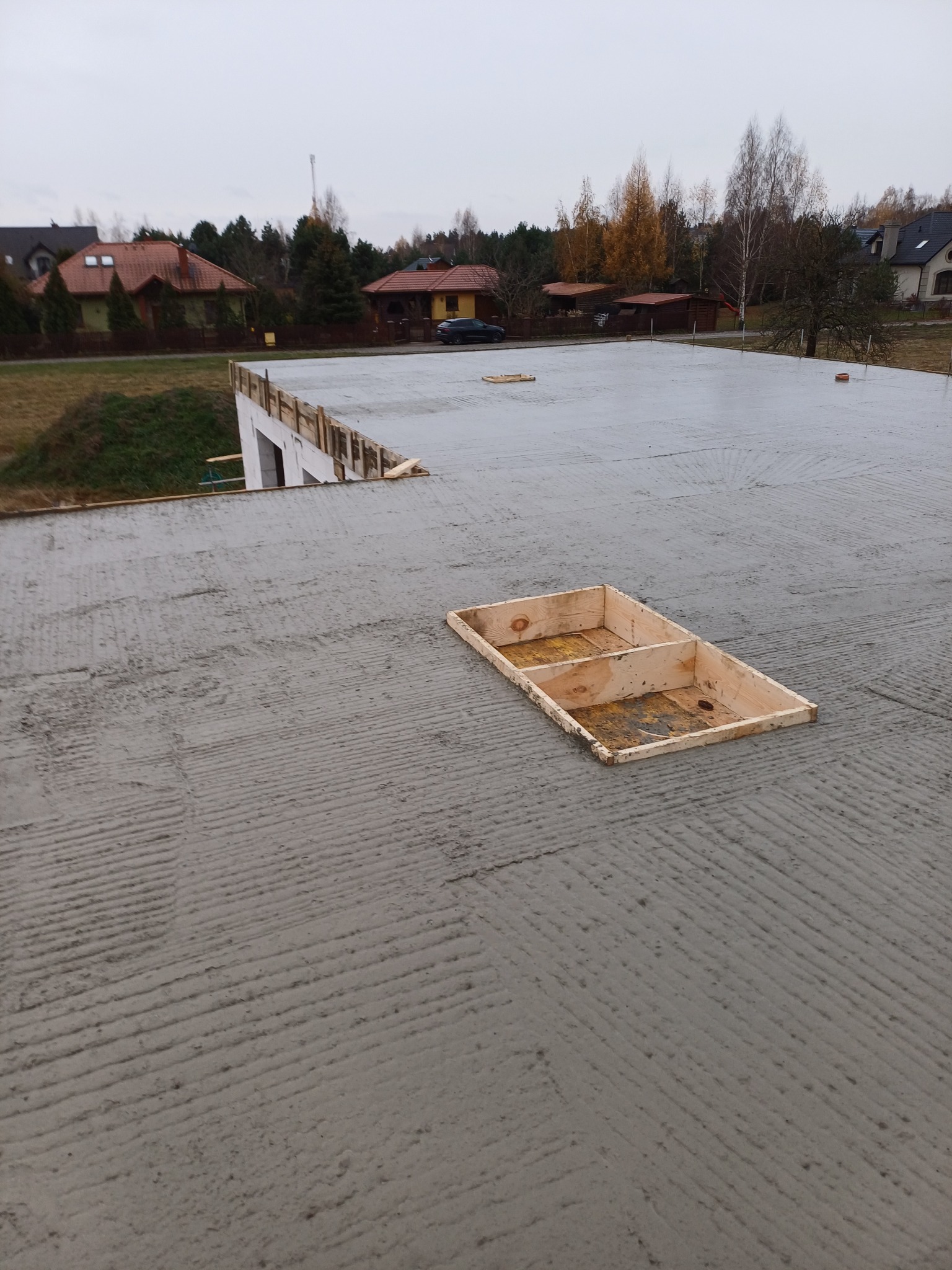 Świeżo wylany betonowy strop z widocznymi śladami po zacieraniu, na którym umieszczono drewnianą formę szalunkową. W tle zabudowa jednorodzinna i pochmurne niebo.