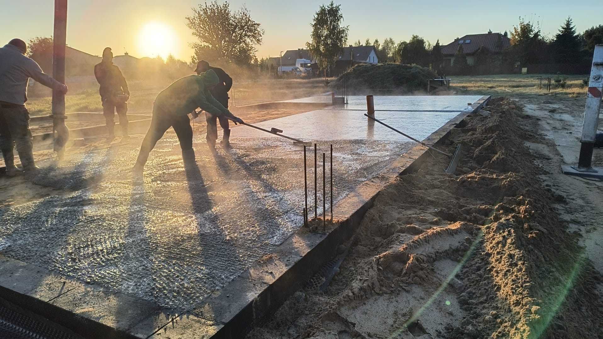 Wylewanie fundamentów domu jednorodzinnego o wschodzie słońca; robotnicy rozprowadzają mokry beton na przygotowanym zbrojeniu i szalunku.