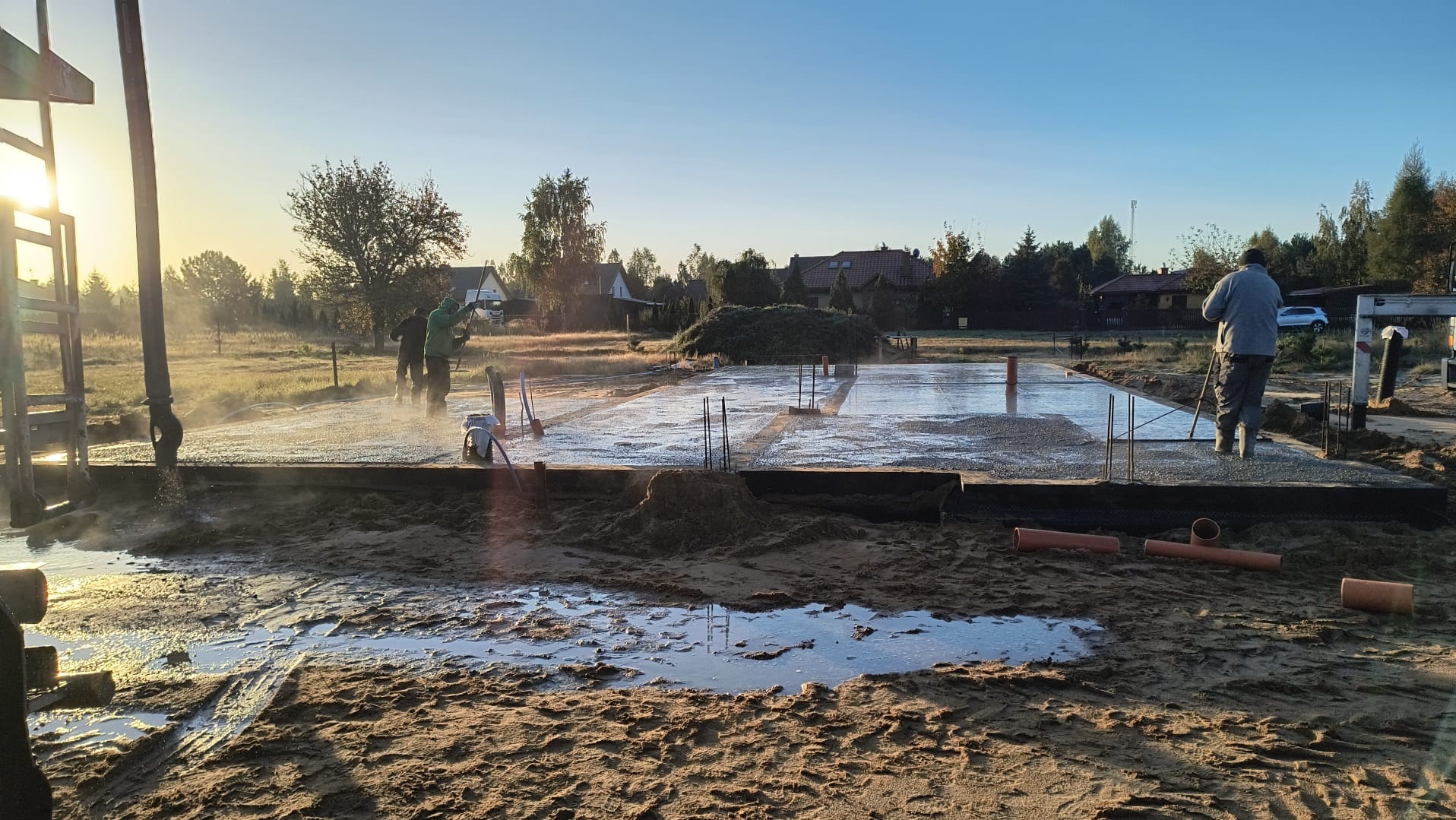 Wylewanie fundamentów pod budowę domu jednorodzinnego, widoczne zbrojenia i pracownicy w trakcie rozprowadzania betonu, w tle zabudowa jednorodzinna i wschodzące słońce.