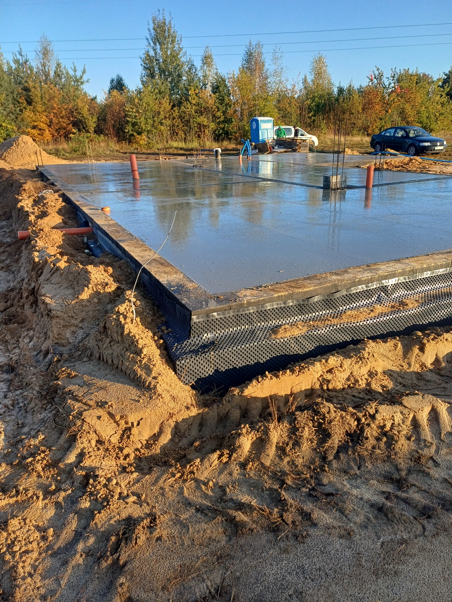 Wylany fundament pod budowę domu, widoczne zbrojenia, szalunki i izolacja pionowa, w tle drzewa i błękitne niebo.