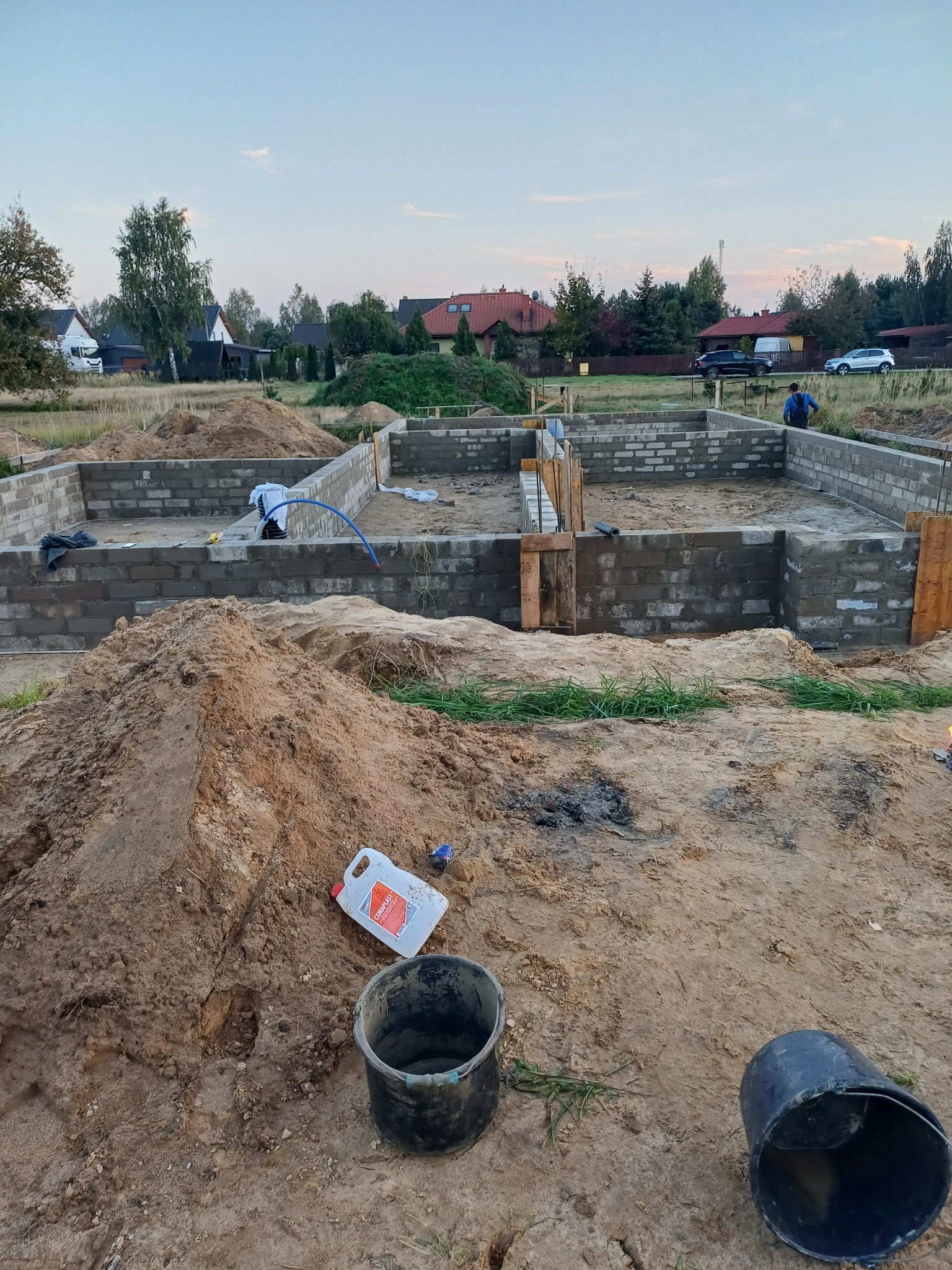 Wykopane fundamenty z bloczków betonowych na placu budowy, widoczne szalunki, hałda piasku, wiadro i kanister z preparatem budowlanym, w tle zabudowa jednorodzinna.