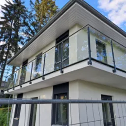 Nowoczesny balkon z balustradą ze szkła i czarnych metalowych słupków, widok z dołu, z tymczasowym ogrodzeniem budowlanym na pierwszym planie i drzewami w tle.