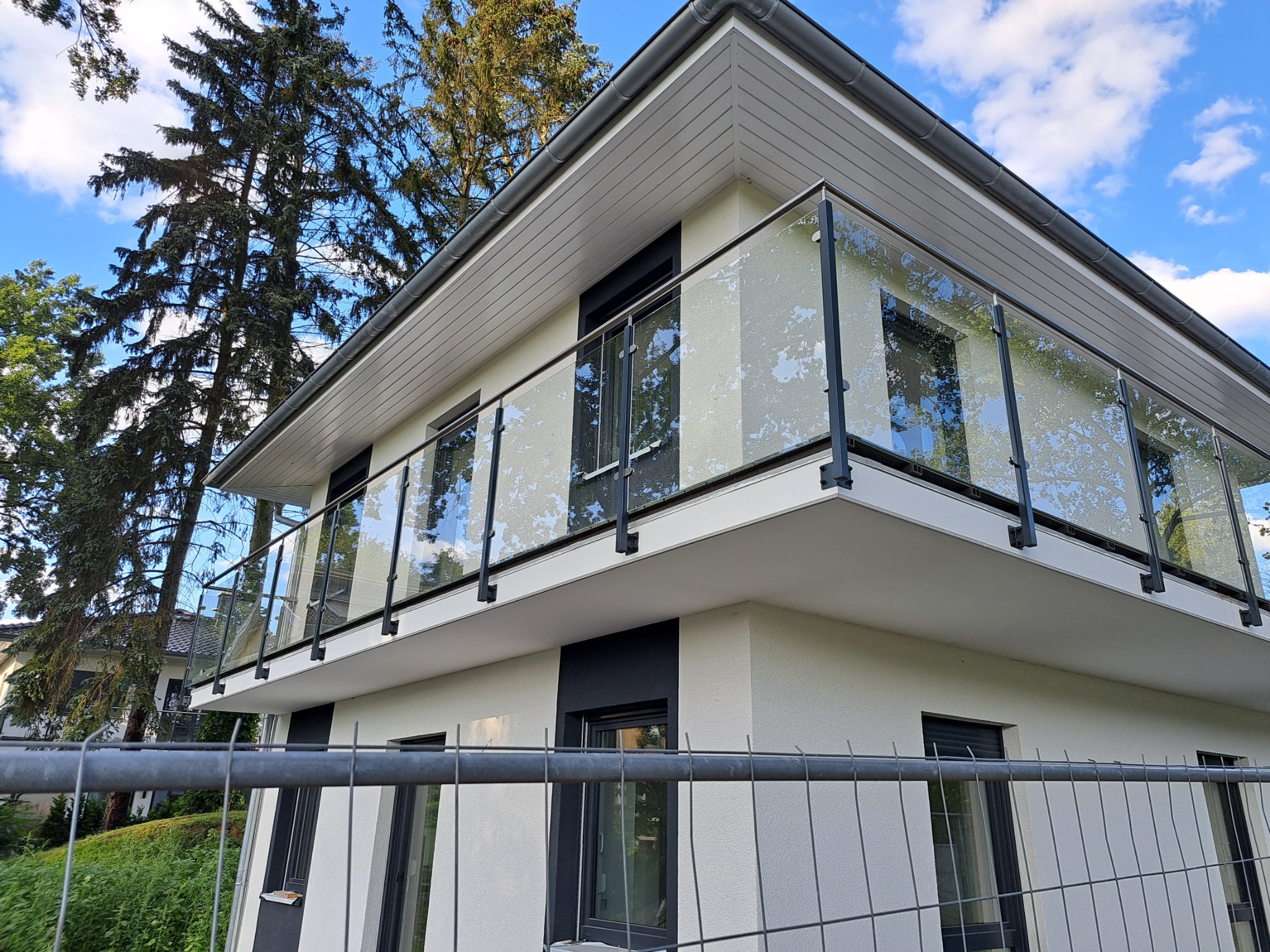 Nowoczesny balkon z balustradą ze szkła i czarnych metalowych słupków, widok z dołu, z tymczasowym ogrodzeniem budowlanym na pierwszym planie i drzewami w tle.