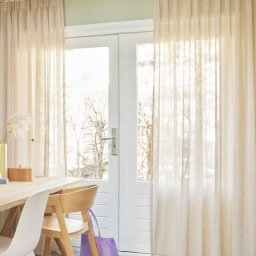 PD Porter Design Paulina Porter - Jasne, delikatne zasłony z lnu, upięte na karniszu nad białymi drzwiami balkonowymi, z widocznym fragmentem drewnianego stołu i krzesła oraz fioletową torbą.