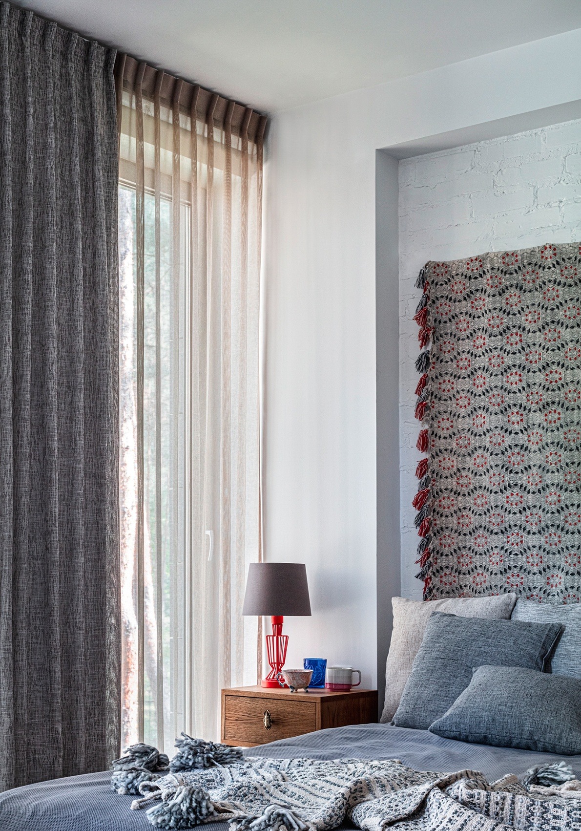 Sypialnia z dwuwarstwowymi zasłonami: szara, gruba zasłona i beżowa, transparentna firana system Flex, drewniana szafka nocna z czerwoną lampką i dekoracyjna narzuta na łóżku.