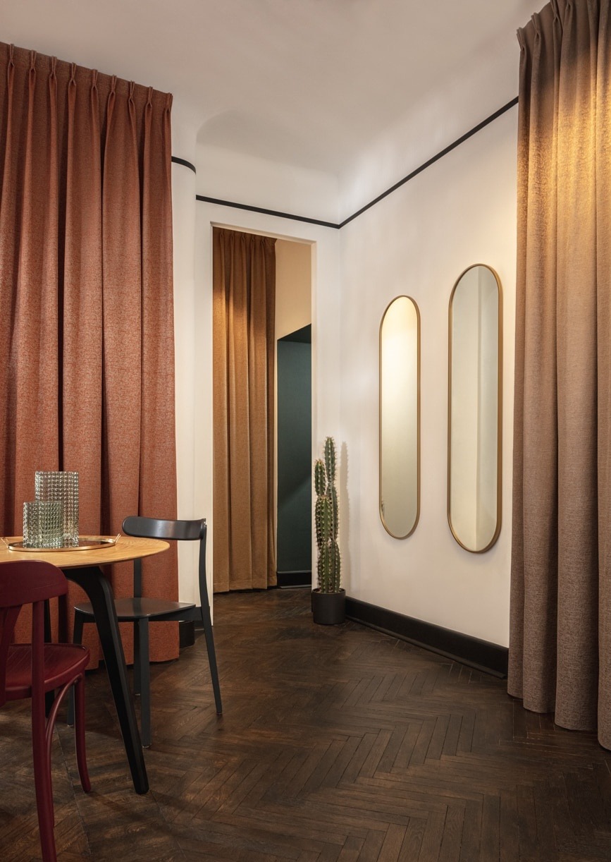 Nowoczesne wnętrze z brązowymi zasłonami w systemie Flex, drewnianym parkietem w jodełkę, okrągłym stołem i dwoma owalnymi lustrami na ścianie.