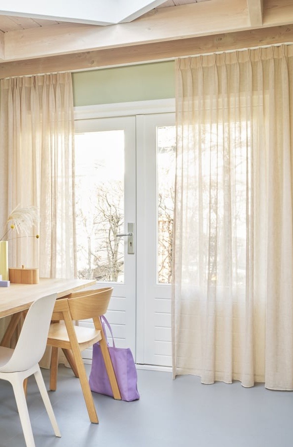 Jasne, delikatne zasłony z lnu, upięte na karniszu nad białymi drzwiami balkonowymi, z widocznym fragmentem drewnianego stołu i krzesła oraz fioletową torbą.
