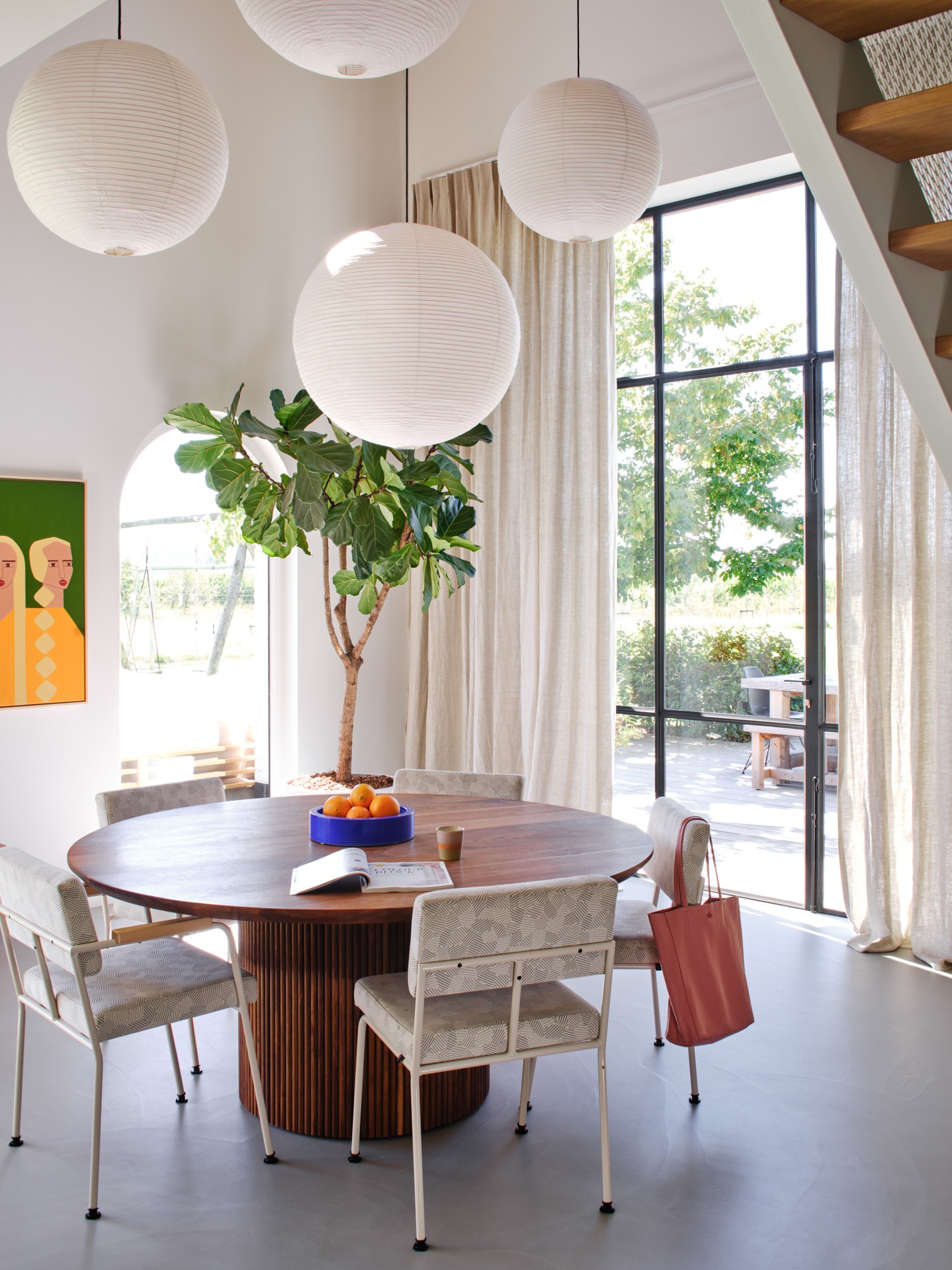 Jasne, przestronne wnętrze z okrągłym drewnianym stołem, nad którym wiszą trzy papierowe lampiony, a w tle widać lniane zasłony przy dużym oknie z czarnymi szprosami, oraz schody.