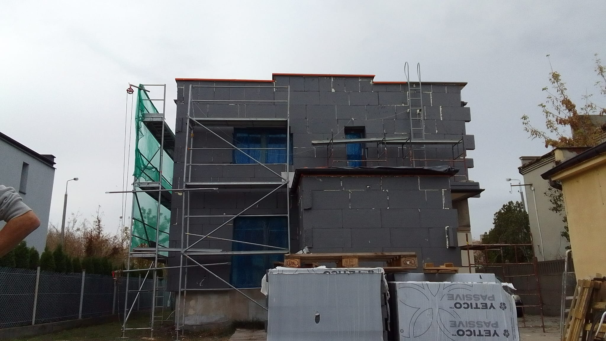 Dom w trakcie ocieplania elewacji styropianem grafitowym, widoczne rusztowania i zabezpieczone okna folią, paleta z materiałem izolacyjnym na pierwszym planie.