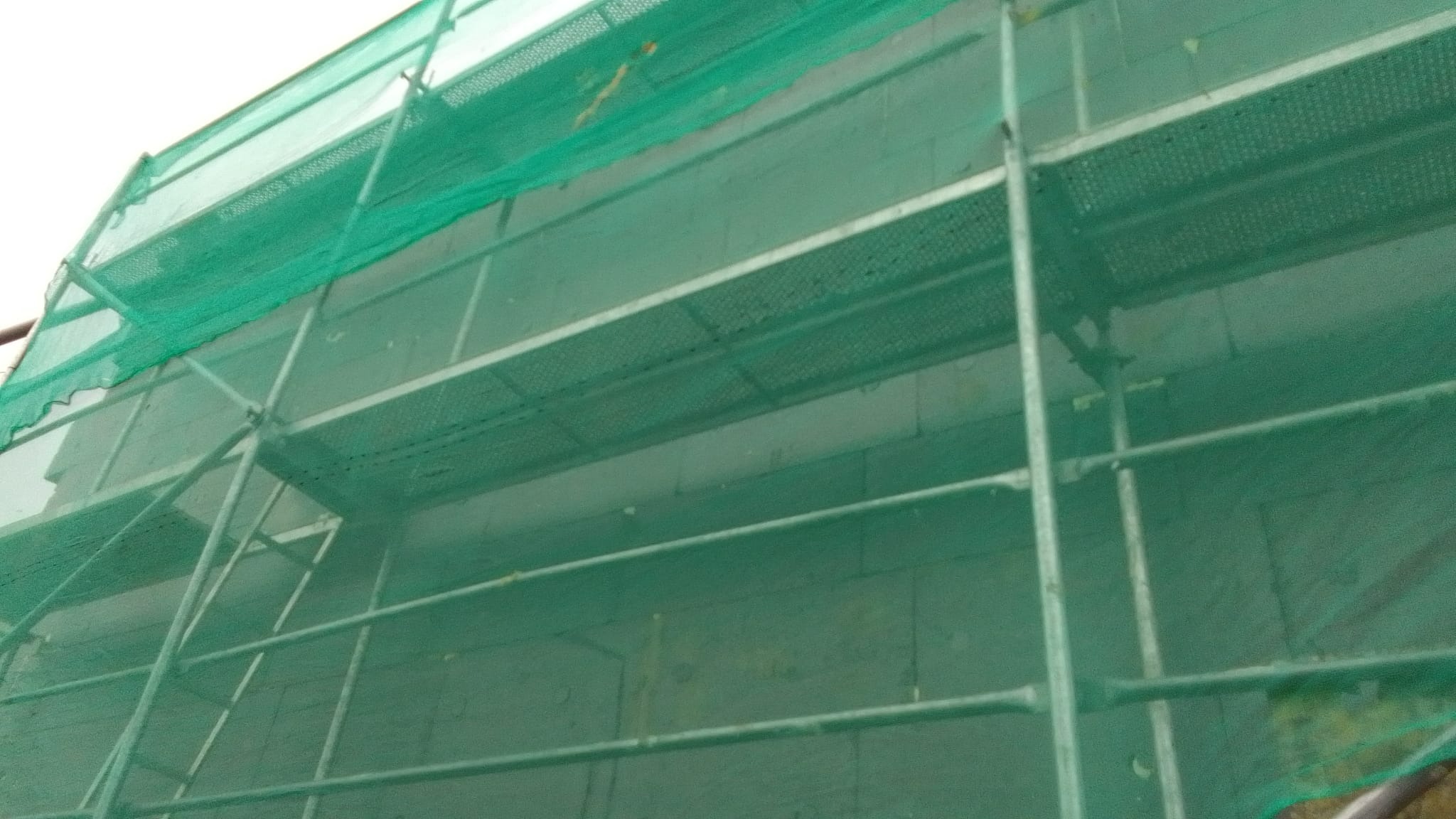 Budynek w trakcie prac elewacyjnych, widoczne rusztowanie z zieloną siatką ochronną i ściana z szarych bloków.