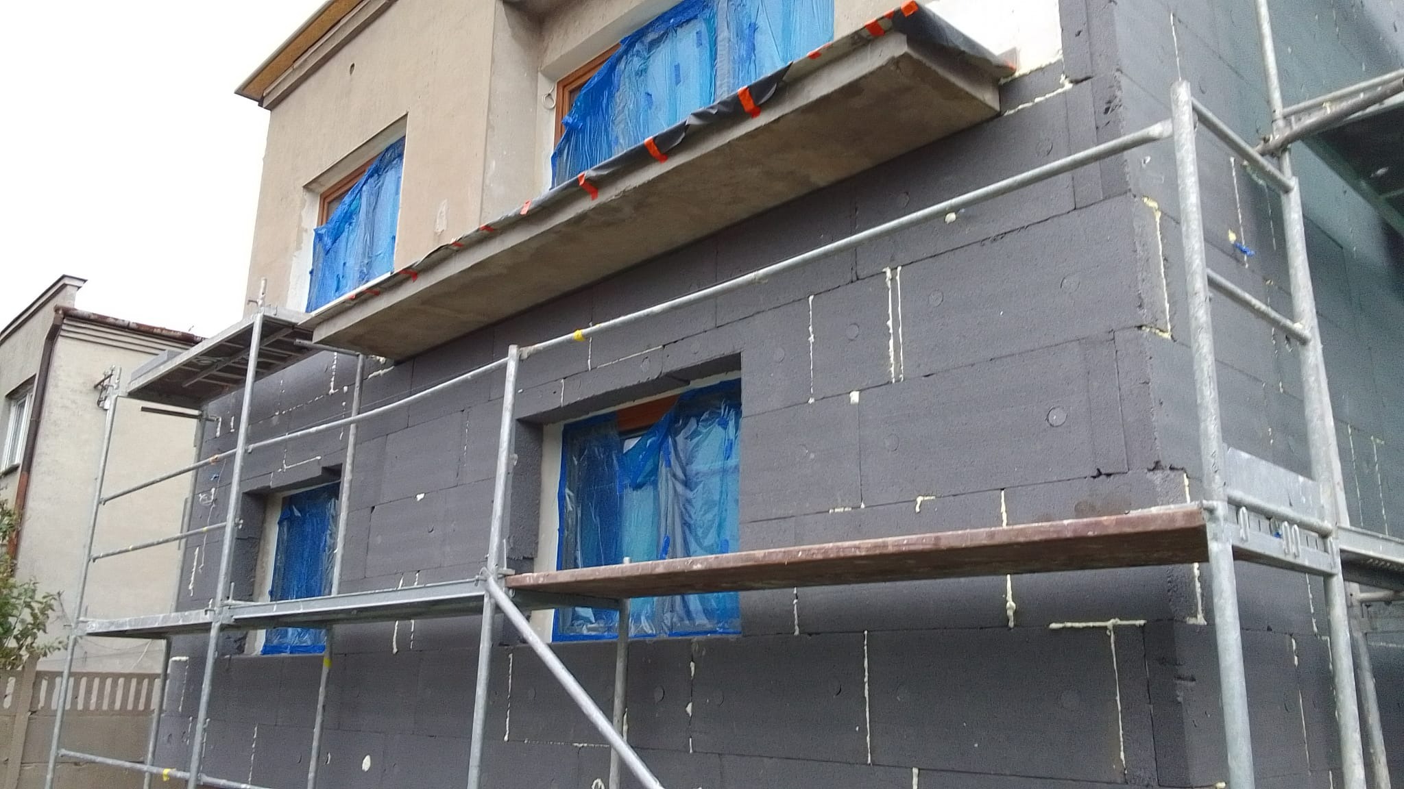 Dom w trakcie prac termoizolacyjnych: szare płyty styropianowe na elewacji, okna zabezpieczone folią, rusztowanie wzdłuż ściany.