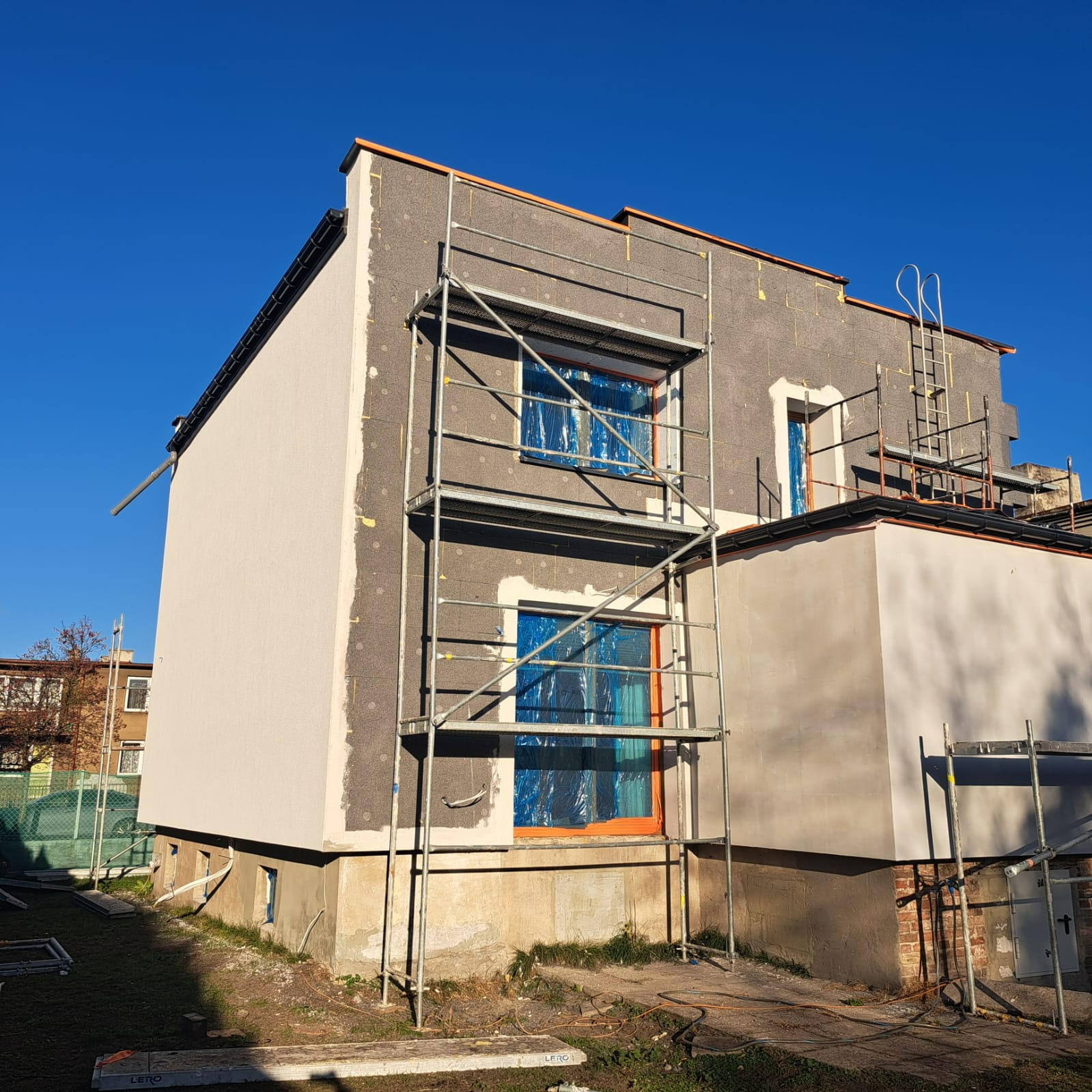 Dwukondygnacyjny budynek w trakcie prac elewacyjnych: jedna ściana otynkowana na jasny kolor, druga z zamontowanym styropianem, okna zabezpieczone folią, rusztowania wzdłuż ścian.
