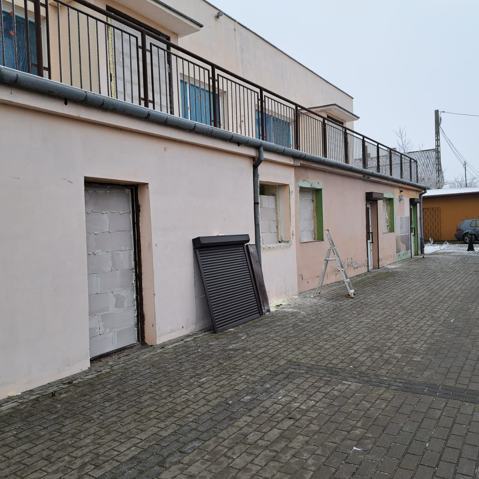 Budynek w trakcie prac elewacyjnych: zabezpieczone okna, częściowo ocieplona fasada, oparta o ścianę roleta i drabina.