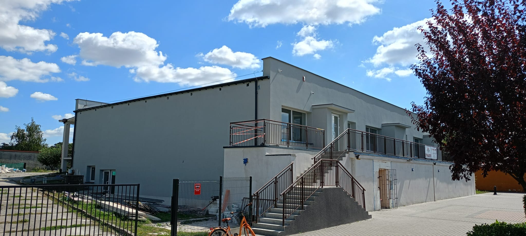 Jasnoszary budynek po remoncie elewacji z nowymi schodami i balustradami, pomarańczowy rower oparty o ogrodzenie, błękitne niebo z białymi chmurami w tle.