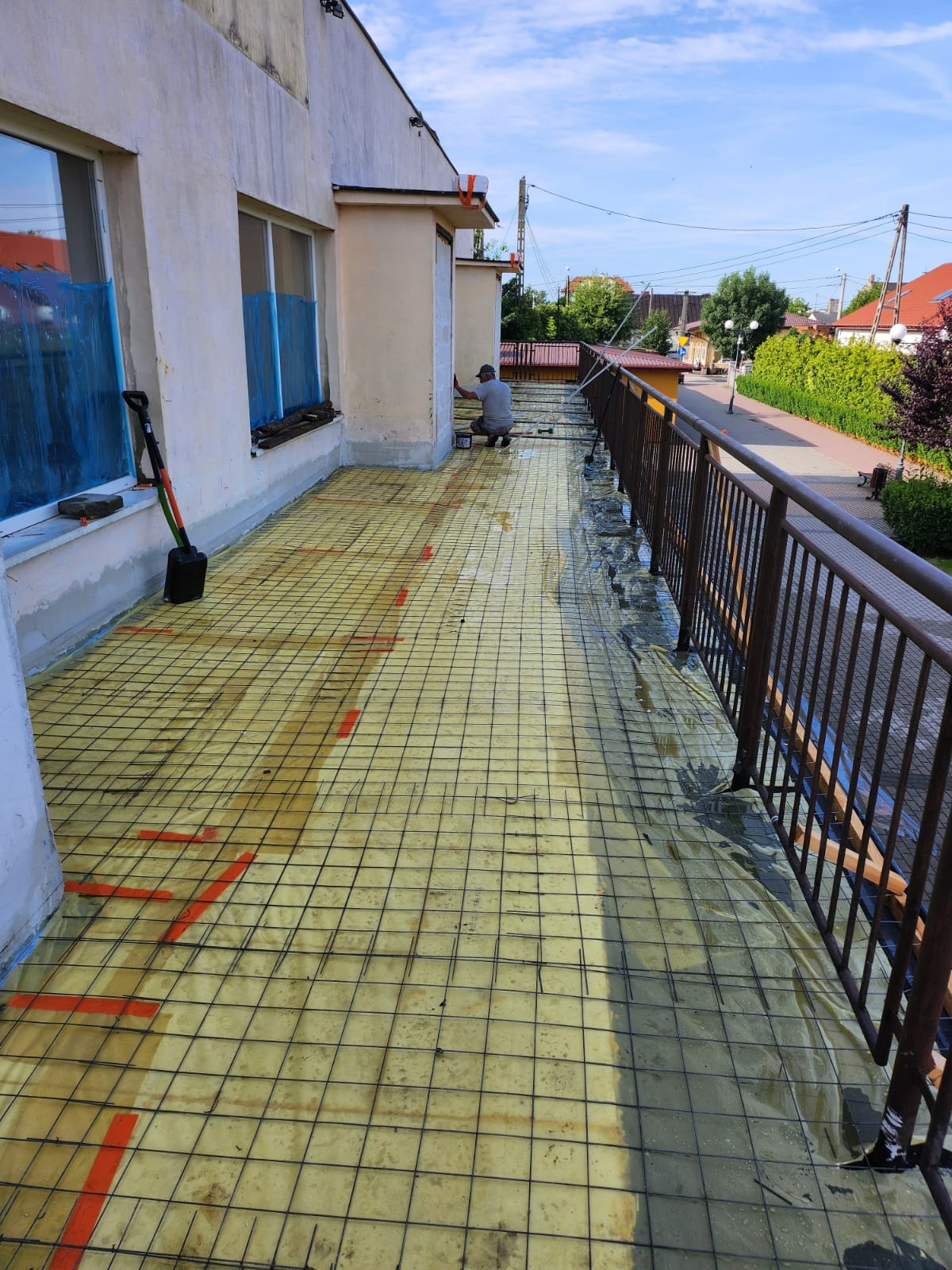 Prace przygotowawcze na balkonie: siatka zbrojeniowa na izolacji, pracownik maluje fragment elewacji budynku. Okna zabezpieczone folią.